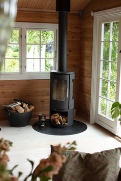 a wood burning stove sitting inside of a living room next to two large windows on the side of a wooden wall