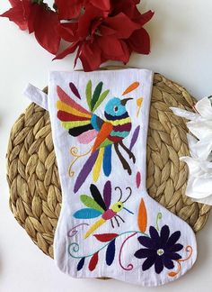 a white christmas stocking with colorful birds on it next to red flowers and wicker basket