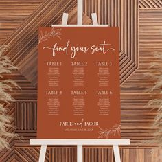 an orange and white wedding seating sign on top of a wooden easel with dried grass