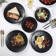 four black plates with food on them sitting on a white table next to utensils