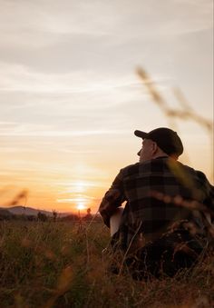 a man sitting in the grass at sunset with his hat on and looking up into the sky