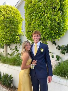 a man and woman in formal wear standing next to each other near some bushes on the side of a building