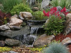 a garden with rocks, flowers and water flowing from it to the ground in front of a pond