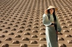 a woman in a long dress and hat standing next to a wall with holes on it