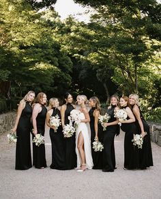 a group of women standing next to each other in front of trees and bushes, all wearing black dresses