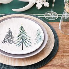 a place setting with pine trees painted on the plates and wine glasses in the background