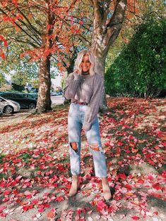 a woman standing in front of a tree with leaves on the ground and wearing ripped jeans