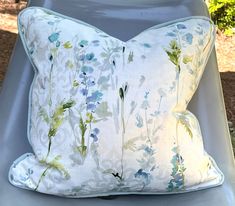 a white pillow with blue and green flowers on it sitting on a chair in the grass