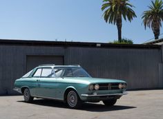 an old green car parked in front of a building with palm trees on the side