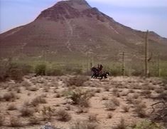 two people riding horses in the desert with a mountain in the backgrouund