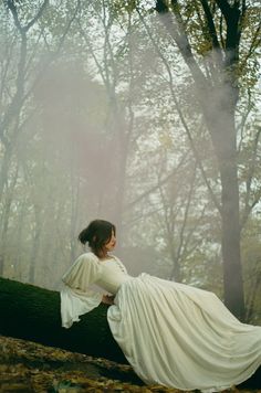 a woman in a long white dress is sitting on a tree trunk with her back to the camera