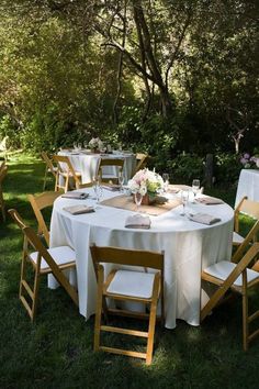 a table set up with white linens and wooden chairs for an outdoor dinner party