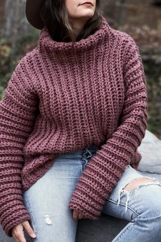 a woman wearing a hat and sweater sitting on a rock with her hands in her pockets