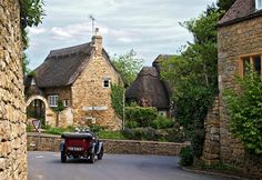 an old car driving down the road in front of a stone building with thatched roof