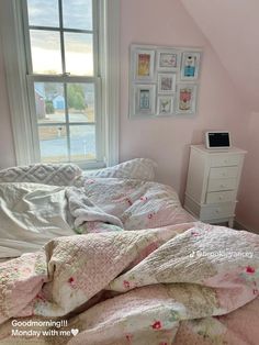 an unmade bed in a pink bedroom with pictures on the wall and window above it