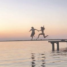 two people jumping off a dock into the water at sunset or dawn with their arms in the air