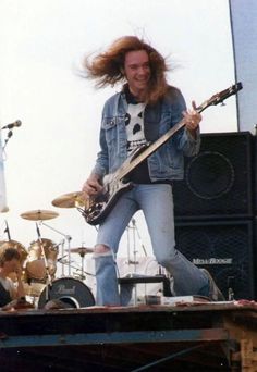 a man with long hair playing guitar on stage at a music festival, in front of other musicians