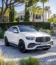 a white mercedes suv is parked in front of a house with palm trees and bushes