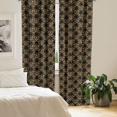 a white bed sitting next to a window covered in black and gold curtain panels on top of a wooden floor