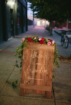 a wooden sign with flowers on it sitting on the side of a street next to a sidewalk