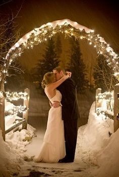 a bride and groom kissing in front of an archway with christmas lights on the trees