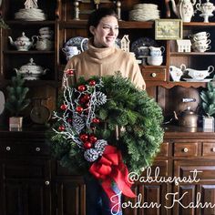 a woman holding a wreath in front of a wooden cabinet with christmas decorations on it