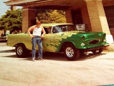 a man standing next to an old green car in front of a gas station with his hands on his hips