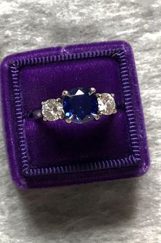 an engagement ring in a purple velvet box with three diamonds on the sides and a blue stone center