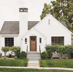 a white house with a brown door and steps leading up to the front door is surrounded by greenery