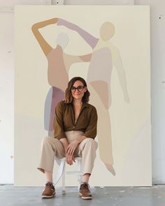 a woman sitting on top of a white chair in front of a wall with an abstract painting behind her