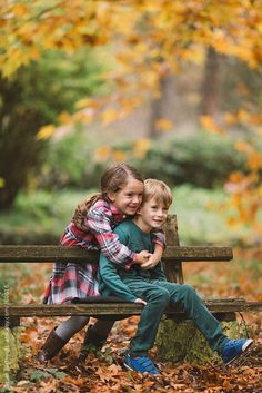 two young children sitting on a bench in the woods with leaves all around them and one child hugging his back