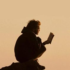 a man sitting on top of a rock while reading a book in front of the sky