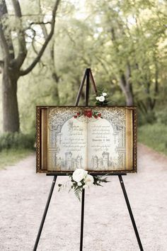 an open book with flowers on it sitting on top of a easel in the middle of a dirt road