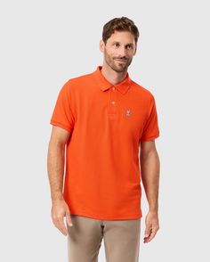 A man with short hair is wearing the MENS CLASSIC PIQUE POLO SHIRT - B6K001CRPC by Psycho Bunny, in orange, paired with beige pants, as he stands against a plain gray background. Classic Polo Shirt With Button Closure, Luxury Workwear Button-up Polo Shirt, Orange Collared Cotton Polo Shirt, Solid Color Button-up Polo Shirt With Placket, Classic Polo Collar T-shirt With Button Closure, Bunny Logo, Pique Polo Shirt, Mother Of Pearl Buttons, Pima Cotton