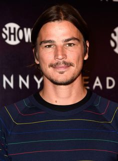 a man with long hair wearing a striped shirt and looking at the camera while standing in front of a black backdrop