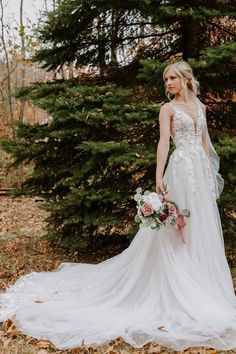 a woman in a white wedding dress standing next to some trees and holding a bouquet