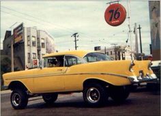 an old yellow car is driving down the street in front of a gas station sign