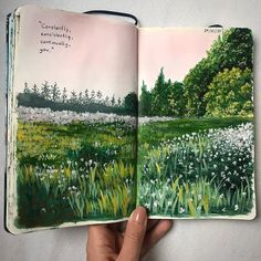 a hand holding an open book with watercolors on the pages and trees in the background