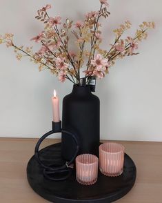 a black vase with pink flowers in it and some candles sitting on a table next to it