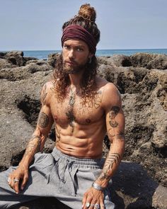 a man with long hair and tattoos sitting on some rocks near the ocean wearing a bandana