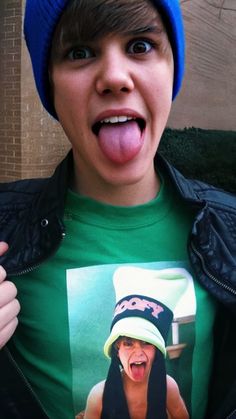 a young man wearing a green shirt and blue hat making a funny face with his tongue out