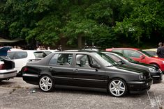 several cars parked in a parking lot with people standing around them and trees behind them