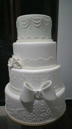 a three tiered white wedding cake with bows and pearls on the top, sitting on a table