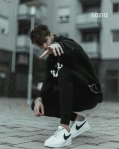 a young man crouching down on the ground pointing at something with his hand while wearing white sneakers