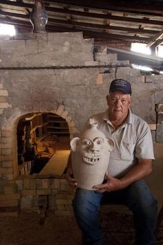 a man sitting in front of a brick oven holding a clay mask with a face on it