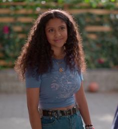 a woman with long curly hair wearing a blue shirt and jeans standing in front of a fence