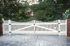 a white picket fence in front of a brick wall and gate with trees behind it