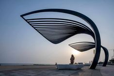 a man standing on top of a white bench next to a large metal sculpture in front of the ocean