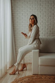 a woman sitting on top of a couch next to a white brick wall holding a cell phone