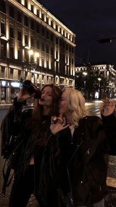 two women standing next to each other in front of a tall building at night time